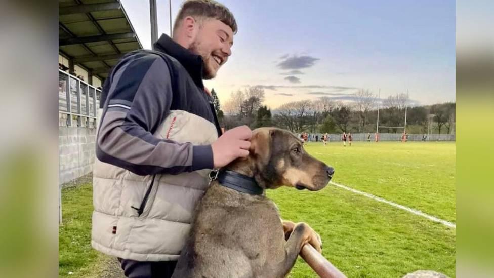 man and foot ball pitch hold dog as they lean on railing