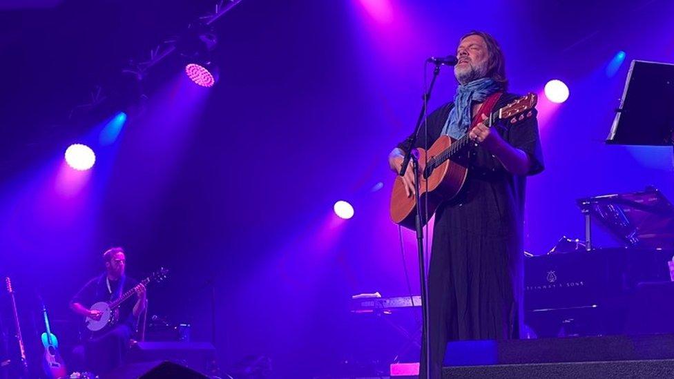 Rufus Wainwright playing the guitar with his eyes closed on stage, bathed in purple light