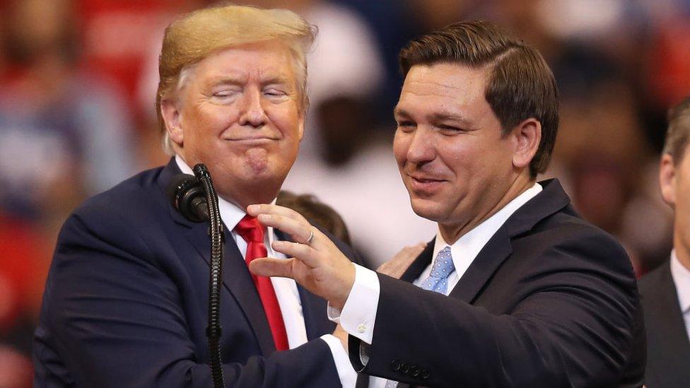 Ron DeSantis appears at a campaign rally with Donald Trump. Both are stood behind a microphone, which Mr DeSantis appears to be reaching towards. Mr Trump is smirking, and Mr DeSantis has a wry smile. The mood is playful and relaxed.