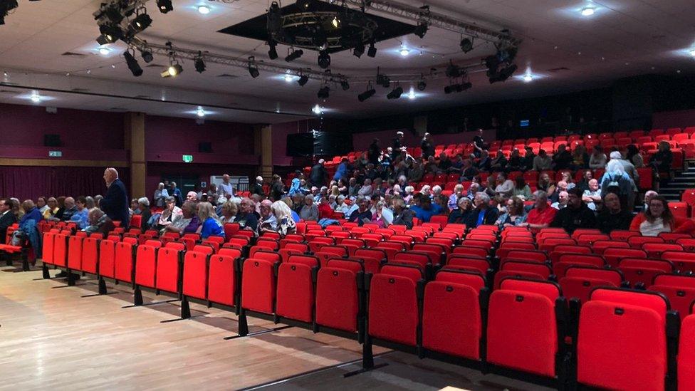 Image of the council meeting. People can be seen sitting in rows of red folding chairs in an auditorium.