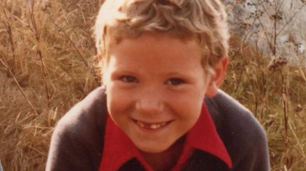 A picture of Hamish, who is stood in front of plants, seemingly leaning in towards the camera. He has short, curly, blonde hair and is wearing a blue jumper and red shirt