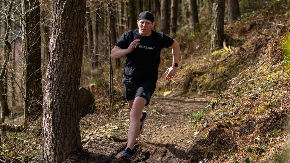 A man in a black hat, t-shirt and shorts is seen running through woodlands. 