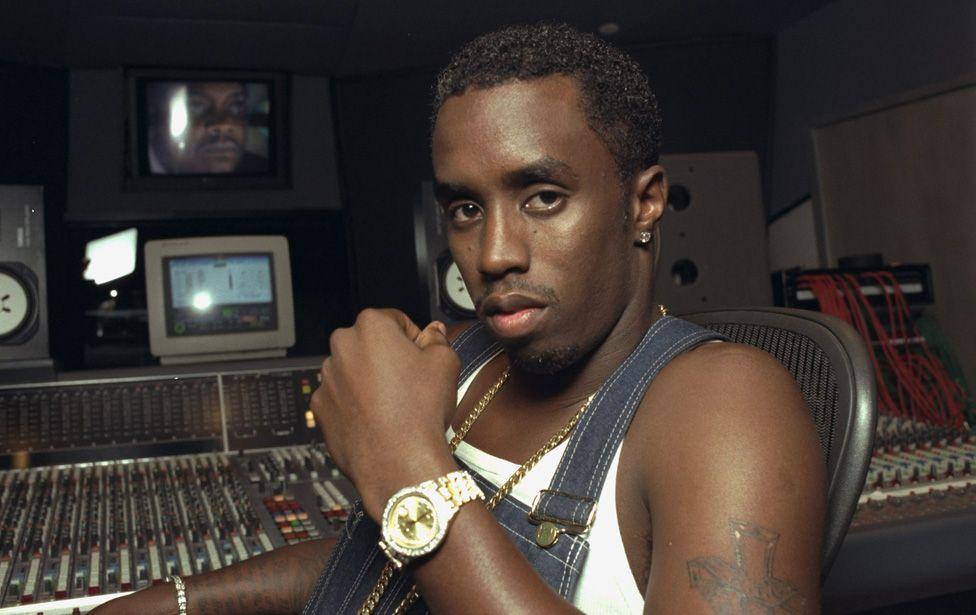 Sean Combs wearing denim dungarees, a white vest and a gold watch, sitting at a mixing desk in a recording studio in 1997