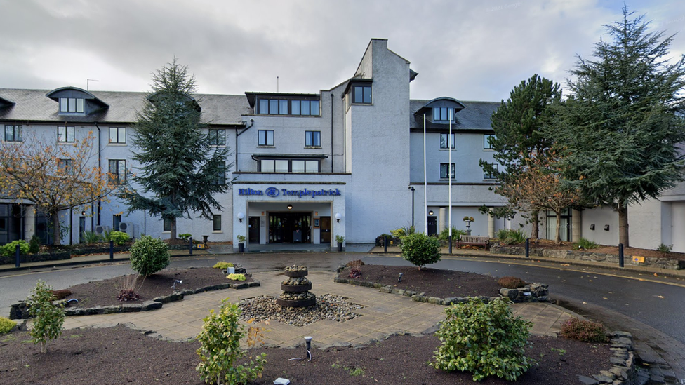 The front of the Hilton Hotel in Templepatrick, with a fountain in the foregound and a small roundabout. The hotel's entrance can be seen under a sign of the hotels name written in blue lettering.