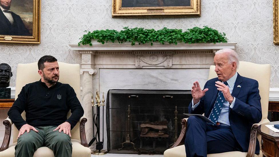 Zelensky listens to Biden as he speaks in the Oval Office. They are both sitting in cream armchairs and a fireplace is in the background with portraits on the wall