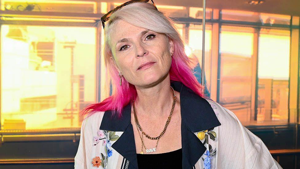 Head and shoulders image of Sister Bliss from Faithless. She has long blonde straight hair which is bright pink on the ends. She has glasses on top of her head and is wearing a floral shirt with black lapels over a black top. She is standing in front of a brightly lit window