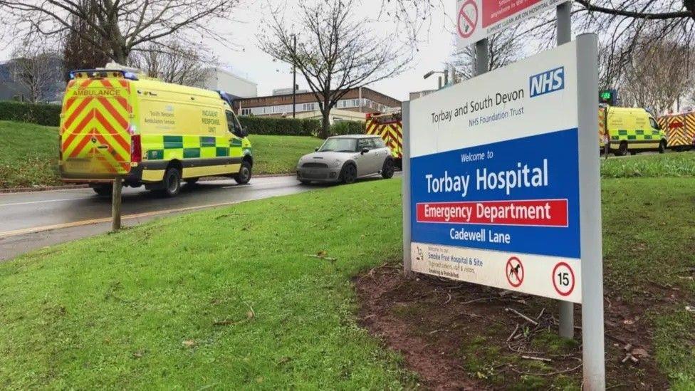 A hospital access road with a sign and a a number of ambulances and cars on the road.