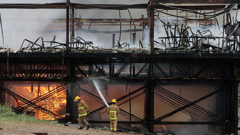 Two firefighters tackle the fire on Fleetwood pier after the blaze destroyed most of the historic pier. One is holding a hose to the burned out shell and one walks past with his head down past an area under the pier that is still ablaze