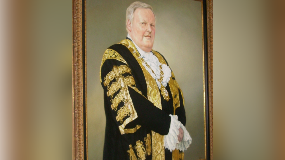 The portrait of Lord Browne is hanging on a wall. He's wearing an official suit. It's black with gold designs and ruffles are peeping out of his sleeves and top.