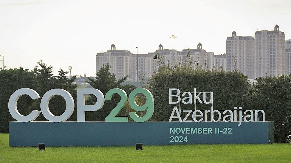 A sign announcing the COP29 United Nations Climate Change Conference November 11-22 2024, against a backdrop of white multi-storey apartment buildings 