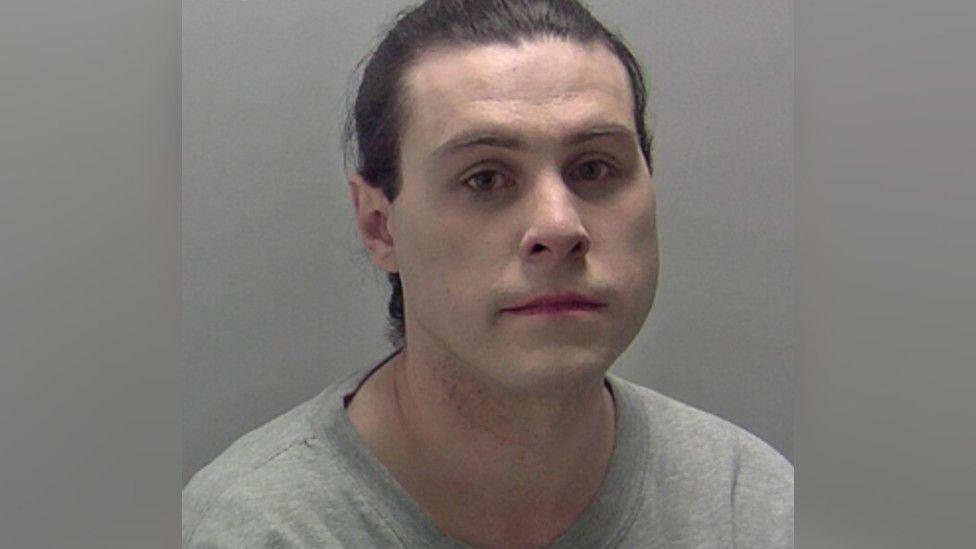 A mugshot of a man who has pale skin and dark hair that is tied back. He is wearing a grey t-shirt.