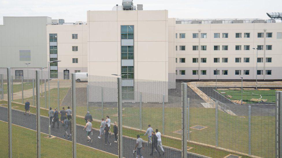 Four-storey concrete prison building with small square windows and one wing at right angles to the main building.  A number of prisoners are walking past on a dark grey path bordered by grass. There is a high see-through prison wall in the foreground.