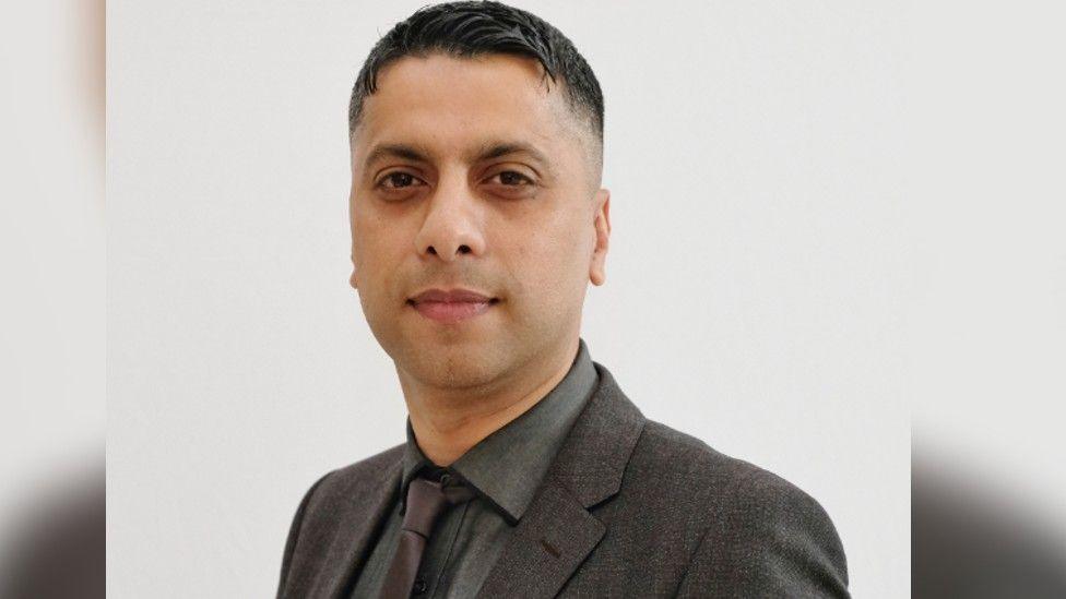  Fiza Matloob smiling at the camera is clean shaven and wearing a grey shirt, brown tie and brown suit jacket.