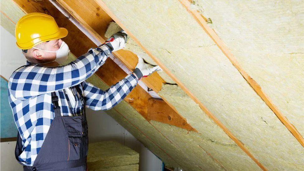 A man in overalls, wearing a mask and a hard hat fitting insulation into a roof