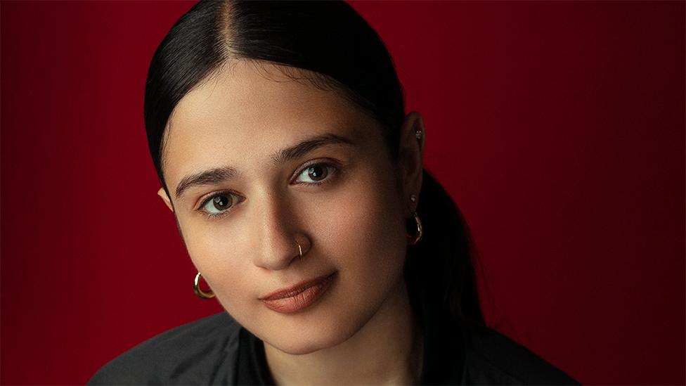 Marva looking at the camera. Her black hair is parted, she is wearing hoop earrings in each ear and has a nose ring. The background is dark red.