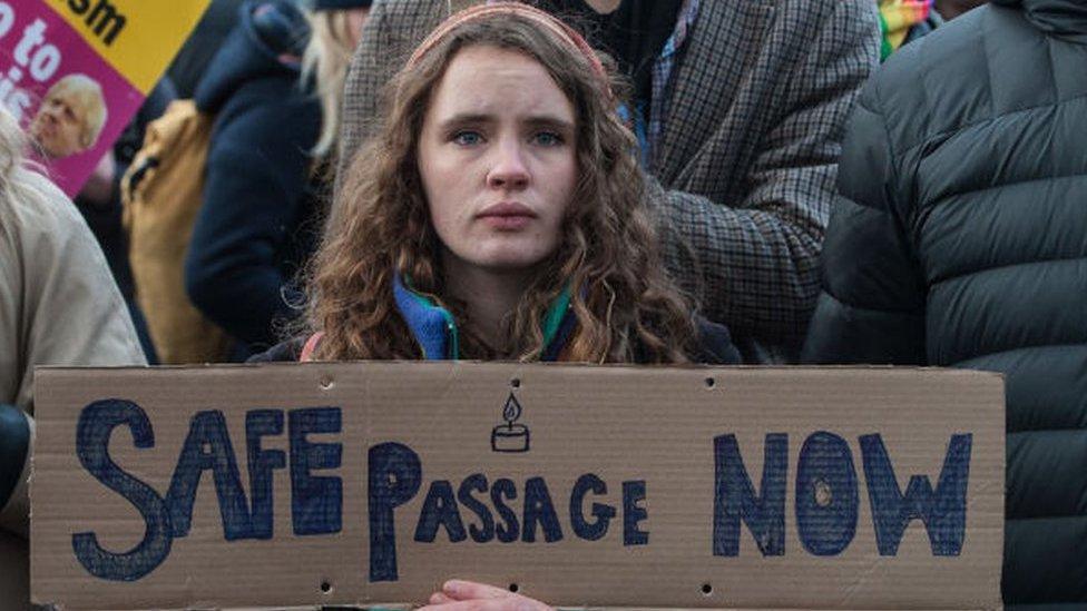 A woman looks sad holding a poster saying safe passage now.