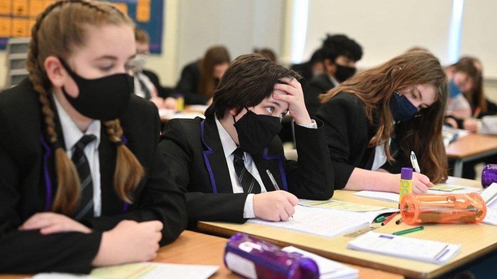 Pupils in black blazers and white shirts with face masks sit at desks with writing materials