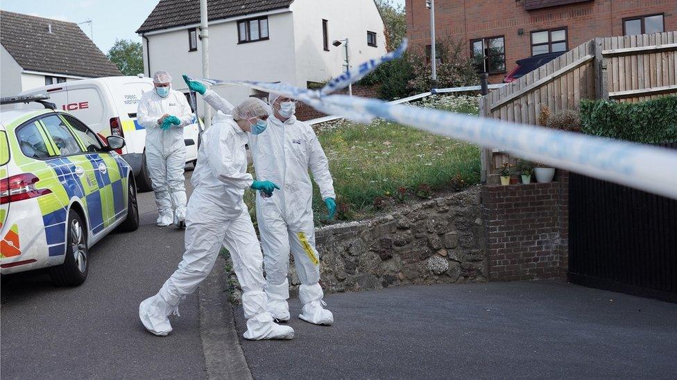 Officers in white protective clothing walk under police tape on driveway