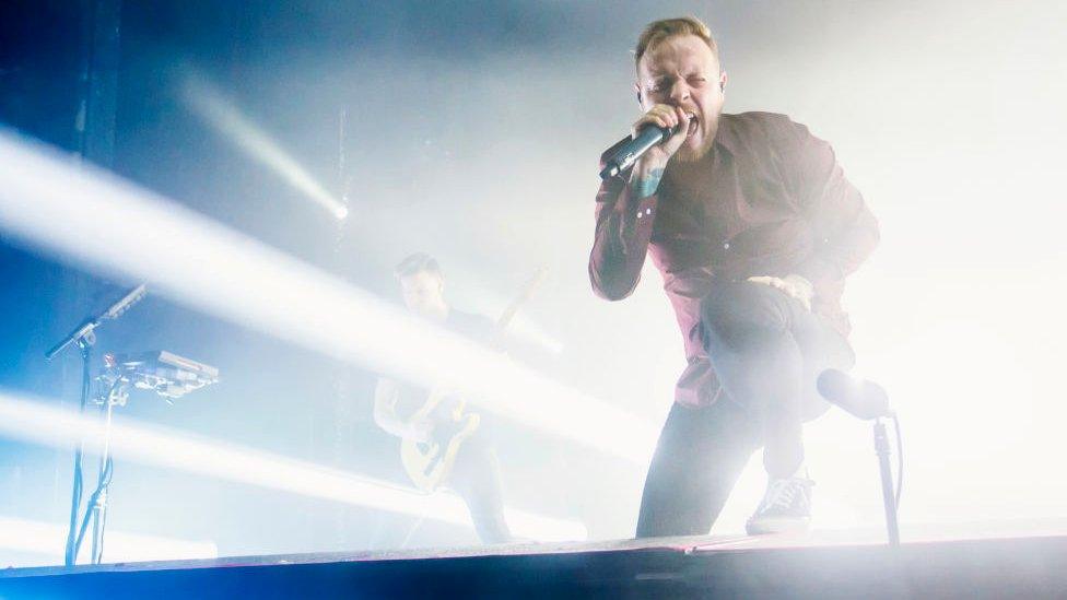 Sam Carter on stage. He's screaming into a microphone, with one leg elevated on a stage monitor in front of him. Bright white stage lights are flooding the space behind him.