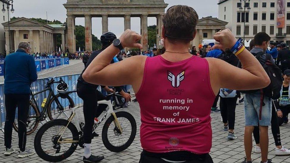 A man dressed in running gear in a crowd with his back to the camera, pointing out the back of his t-shirt, which shows he's running in memory of his son