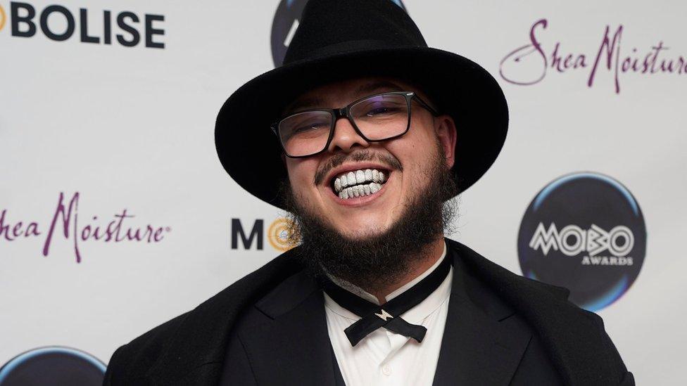 Potter Payper at the Mobo Awards in Sheffield. Potter wears a black fedora hat, a black blazer and a thin black bow tie over a white shirt. He smiles at the camera, revealing a gem-encrusted grill. He wears black rimmed glasses and has a dark brown beard and moustache