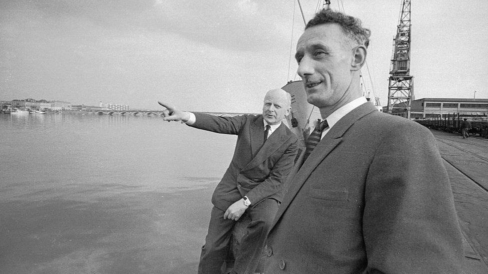 A black and white photograph showing two men - Maj Herbert "Blondie" Hasler and Cpl Bill Sparks - in suits, standing on a dockside with water spanning to the left and a bridge in the background.