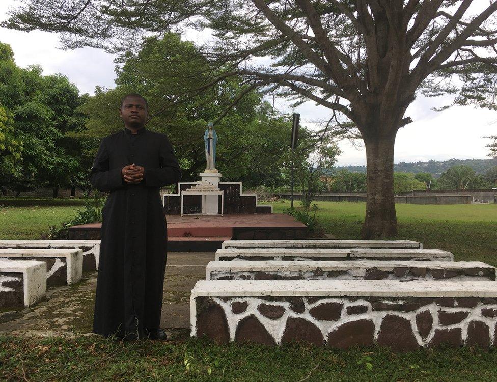 Father Joseph Musubao wears his black cassock and stands close to an outdoor altar and pews