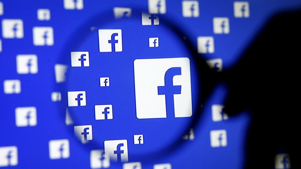 A man poses with a magnifier in front of a Facebook logo