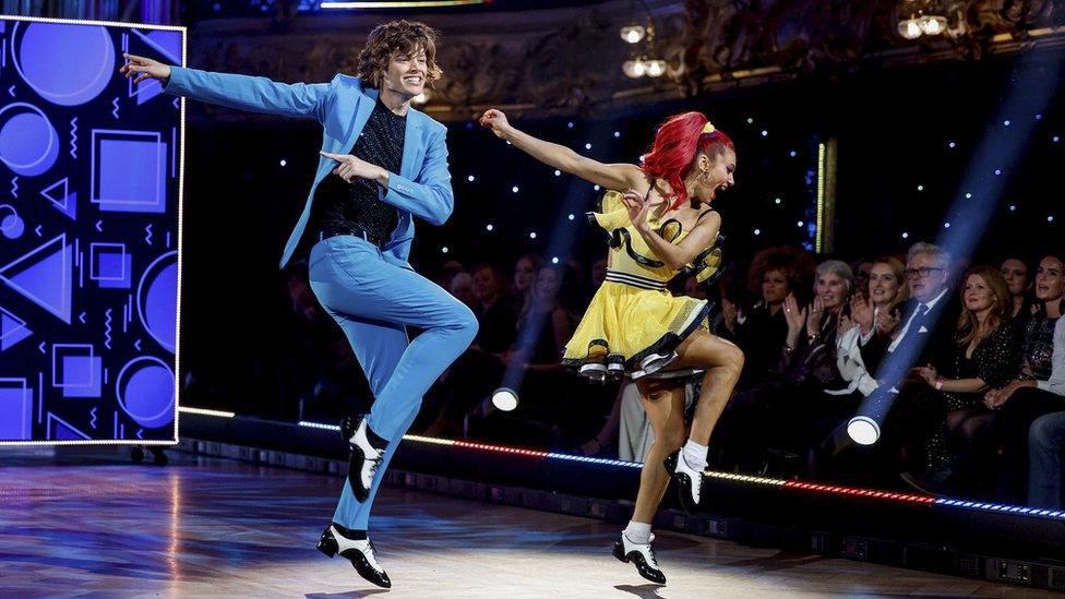 Bobby Brazier and Dianne Buswell during one of their routines. Bobby wears a blue suit over a black shirt, paired with white and black brogue shoes. Dianne, with her red hair tied back in a pony tail, wears a short yellow ruffled dress with a black trim and shoes to match Bobby's. They are pictured hopping with their right legs raised while their right arms swing out. Behind them is the live studio audience and the staging is set with purple shapes on a black screen.