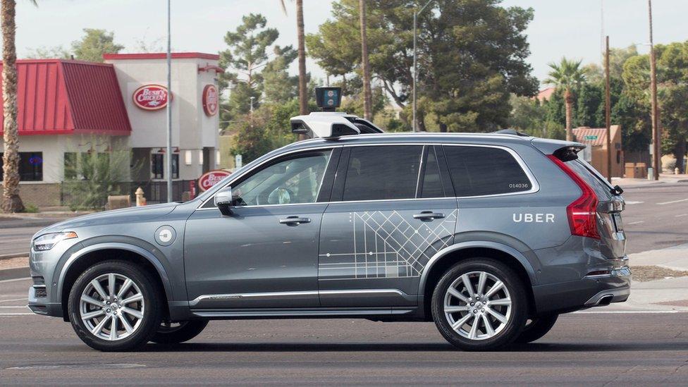 A mettalic grey Volvo car, wrapped with some occasional Uber branding in white vinyl, is seen here with a large mount on top of the vehicle which houses self-driving equipment