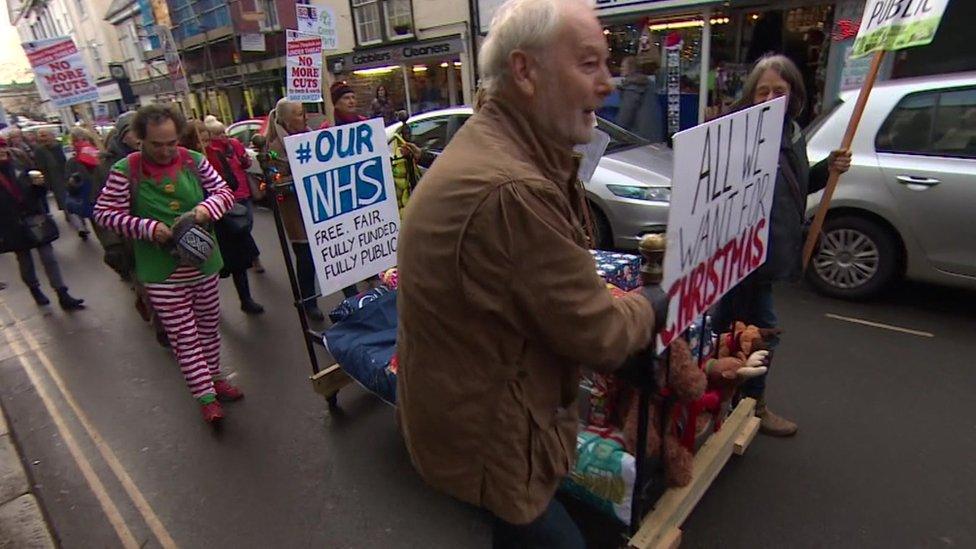 Protesters wheel a hospital bed through Totnes with placards