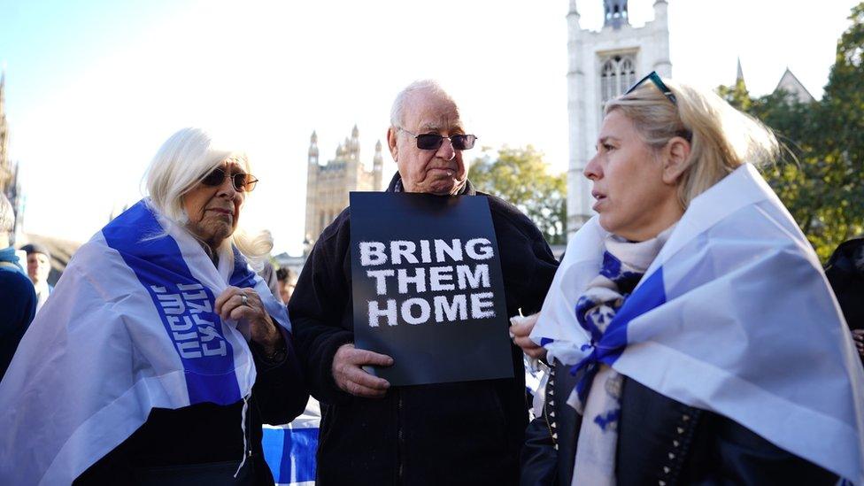 Man holding "Bring them home" sign with two women draped in Israeli flags