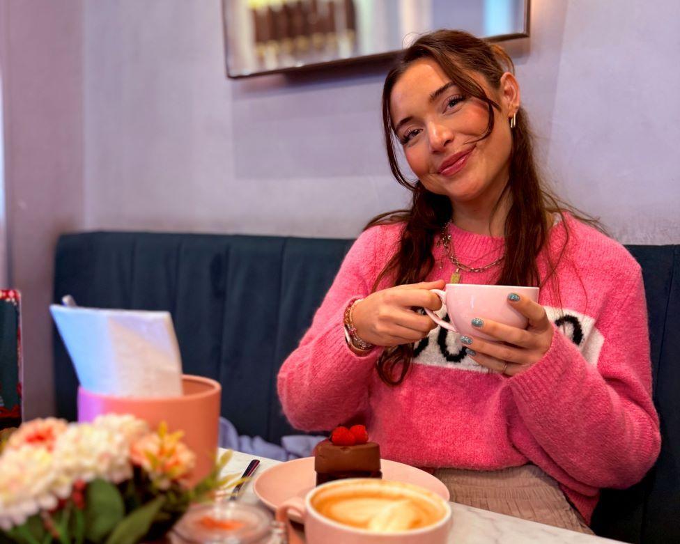 A woman with long brown hair in a bright pink jumper sits in a cafe at a table with a cup of tea, a slice of cake and pot of flowers