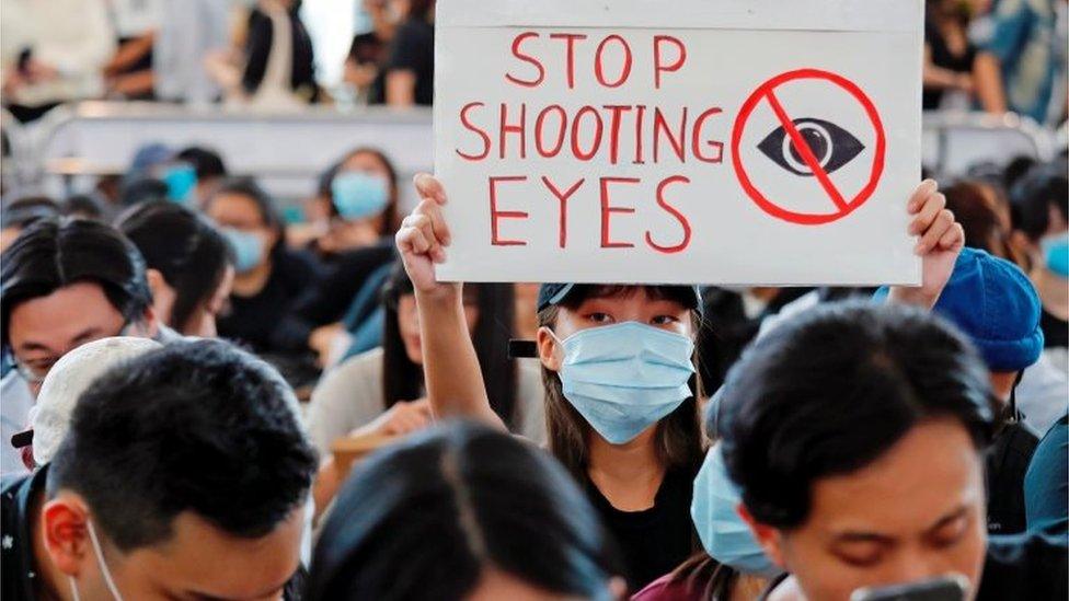 A protester in the airport holds a sign that says "stop shooting eyes"