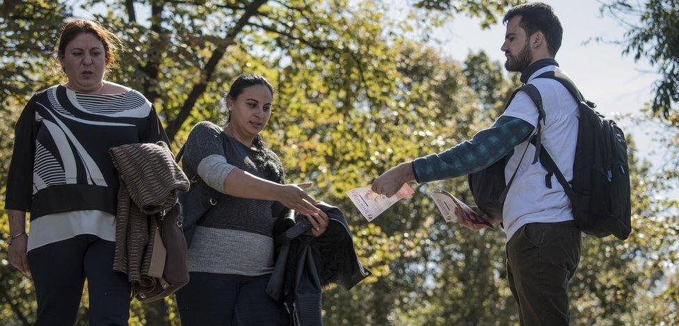 Two women walk past a man in a sunny street handing out flyers - one accepts the offer as she walks past