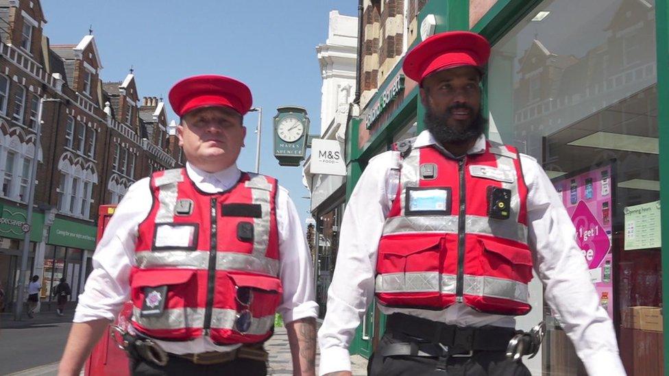 Two Bobbies, wearing red vests, red hats and white shirts, patrol on St John's Road in London.