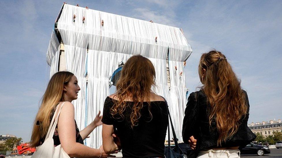 Bystanders look on as workers arrange silver blue fabric, part of the process of wrapping the Arc de Triomphe