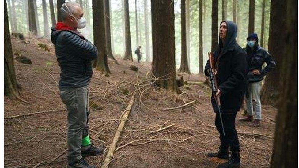 A crew member wears a mask, while an actor holds a fake rifle, surrounded by forest