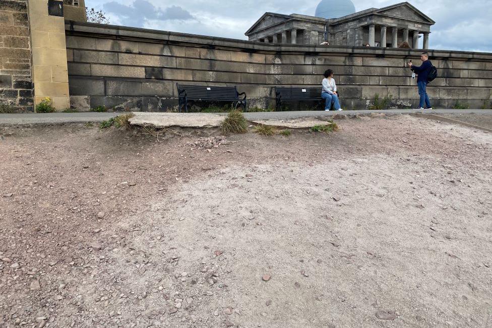 An incline of brown, exposed earth on Calton Hill. A man is taking a photograph of a woman sitting on the bench in the background. 
