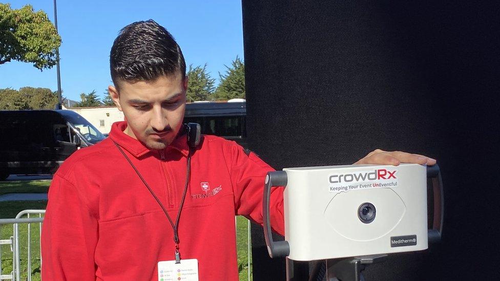 A man wearing a red hoodie looks at a screen with one hand resting causally atop an infrared temperature camera, to scan audience members