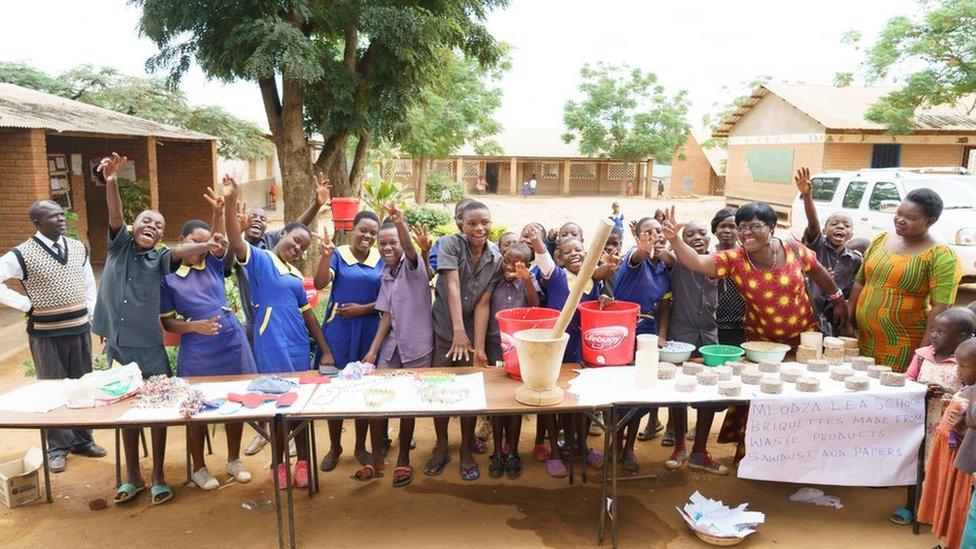 A class of children - both girls and boys - wave at the camera from a table lines with bowls and materials at Mloza primary school in Lilongwe.