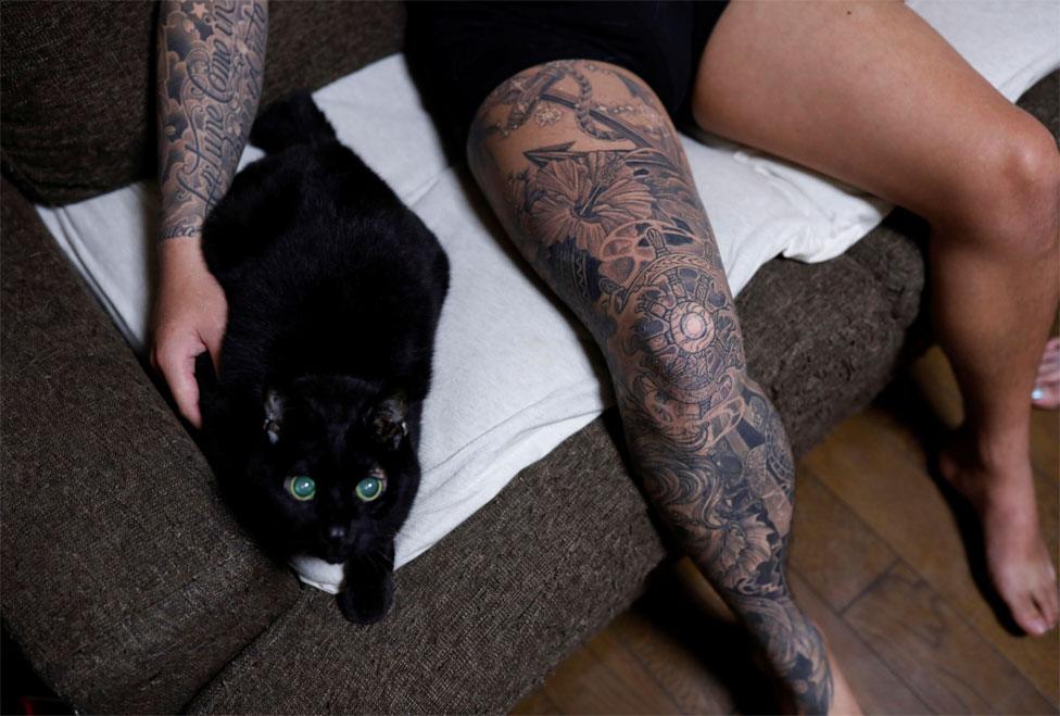 A man with tattoos on his arm and leg pets a black cat on a sofa