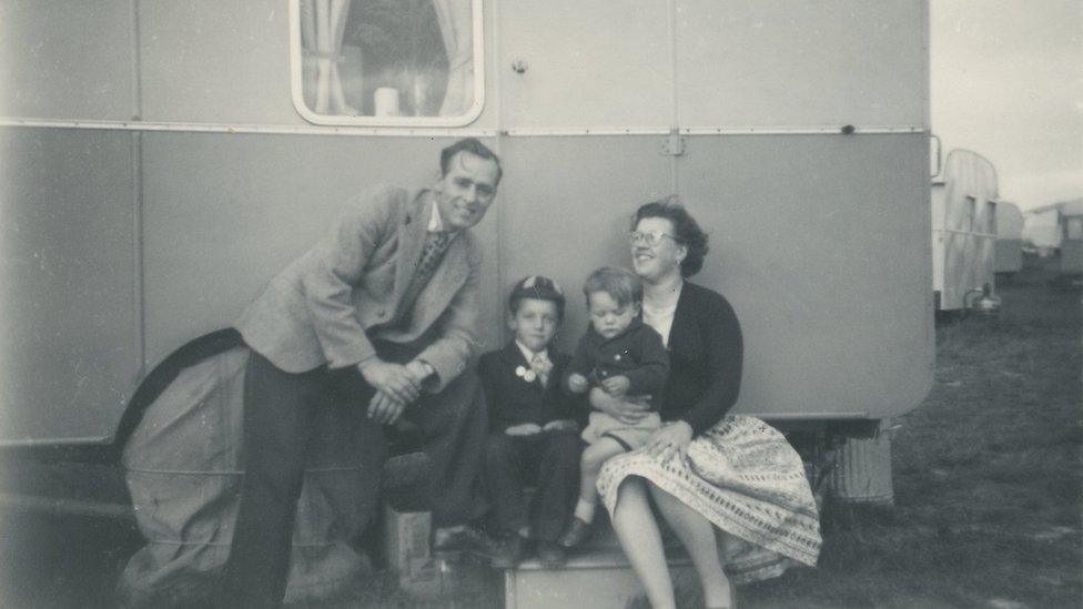 Black and white photo of mother, father and two sons (one wearing a cap) outside a caravan