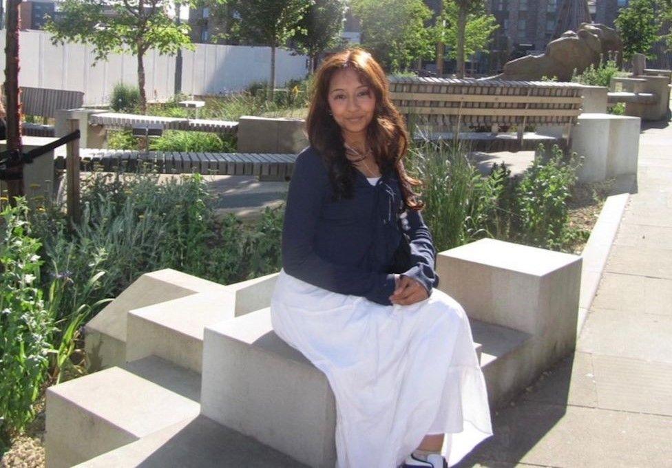 A woman in a navy cardigan and long white skirt sits outside on a modern-looking concrete bench in front of some plants. She is smiling at the camera