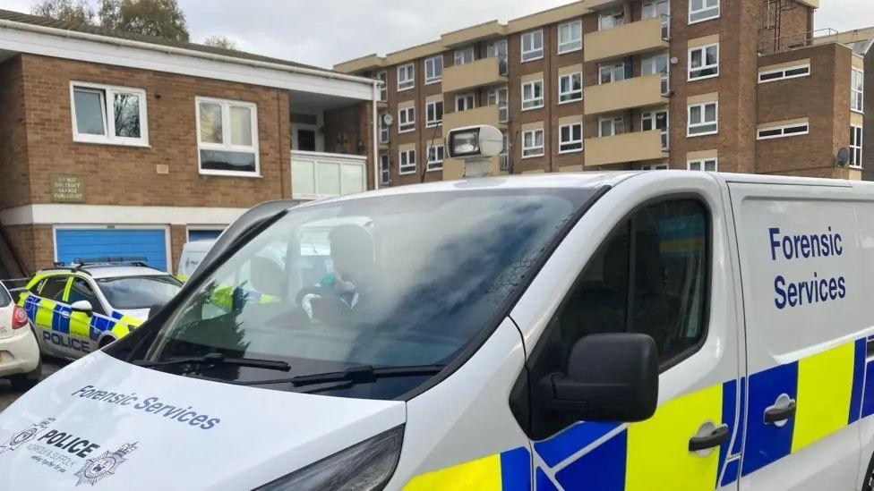 A police van with "Forensic Services" written on the side is parked outside blocks of flats in Mousehold Street in Norwich. Behind the van, on the left of the picture, two other police vehicles can be seen parked in front of a two-storey block.