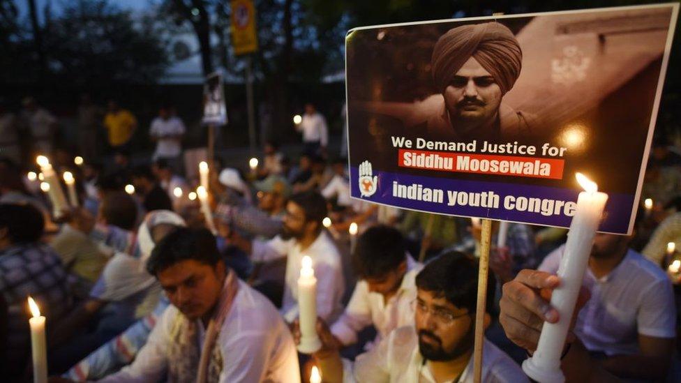 Protesters hold candles at a peaceful protest, most are dressed in white in the twilight scene. A placard in the foreground, showing a black and white picture of Sidhu Moose Wala can be seen. It reads: "We demand justice for Sidhu Moosewala", with the subtitle Indian Youth Congress.