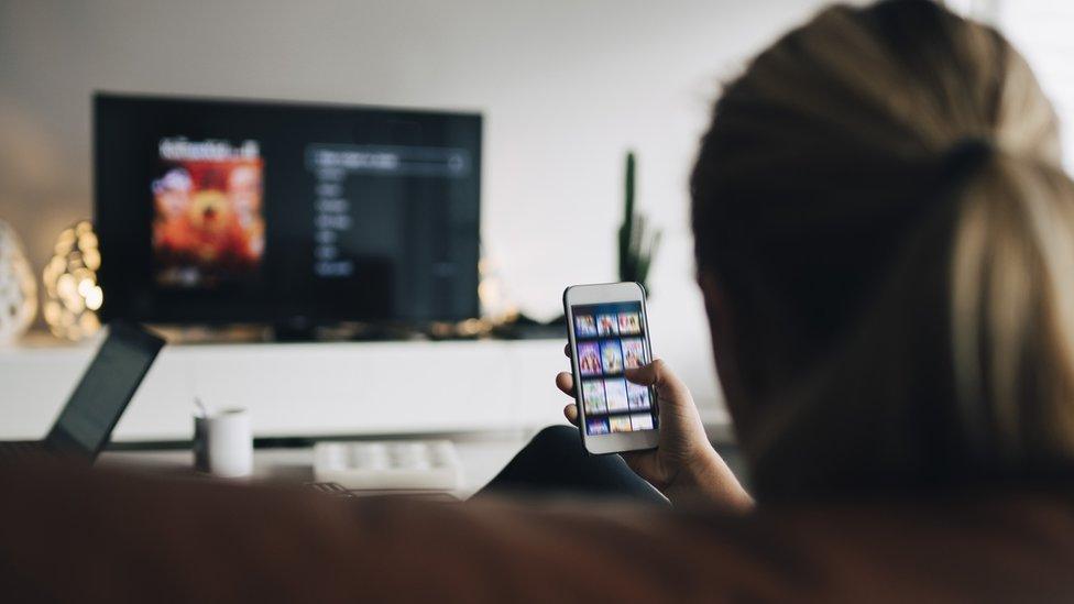 A woman looks at her phone while the TV is on in the background