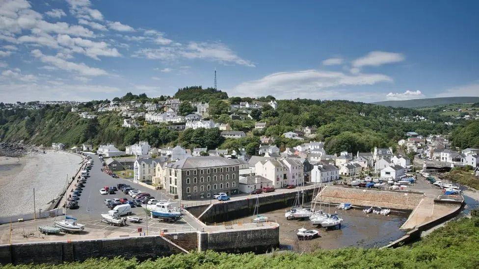 A seaside village with a river and harbour in the foreground, and a hill populated by trees and houses in the background. 