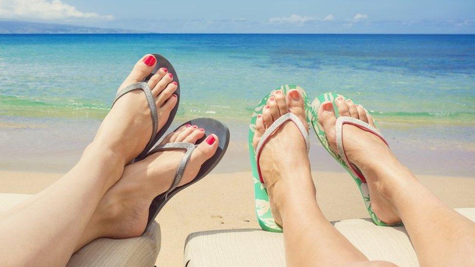 Two women lie next to one another on sun loungers wearing flip flops with the ocean in the background