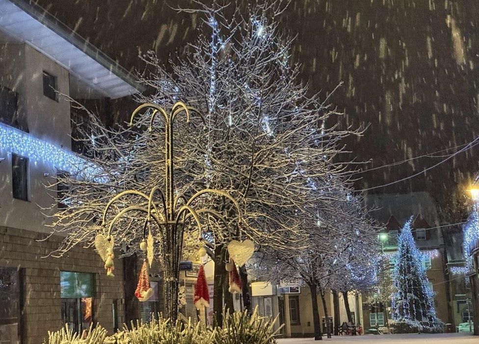 Snow falling in Penicuik, Scotland during a cold snap in the UK. A tree can be seen in the foreground, with its branches covered in snow. There is a Christmas tree and decorations also visible in the picture, with fairy lights being hung from surrounding buildings